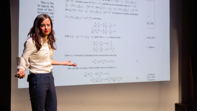 A confused-looking woman stands in front of a projector screen covered in complicated math